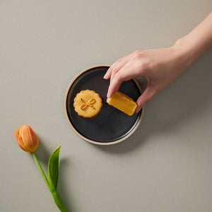 Hands holding a piece of salted egg custard mooncakes from a ceramic plate with another salted egg custard mooncake beside it and a flower on the table