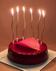 6 lit curvy candles inserted in a pink Raspberry Valrhona Jivara Mousse cake with a pink circular decoration.  
