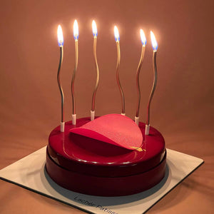 6 lit curvy candles inserted in a pink Raspberry Valrhona Jivara Mousse cake with a pink circular decoration.  